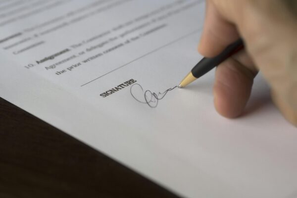 A hand signs a formal contract with a pen on a wooden desk.